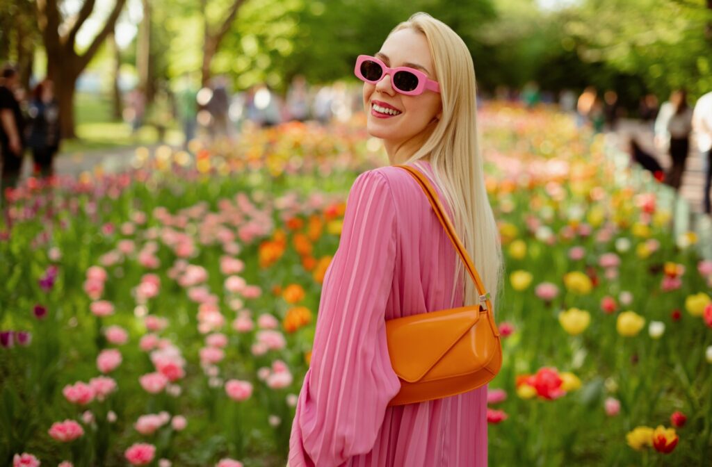 A fashionable young person wearing pink prescription sunglasses stands smiling in front of a field of flowers